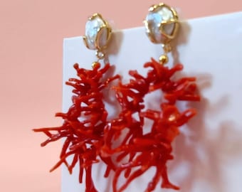 Puede incluir: Un par de pendientes inspirados en el coral. Cada pendiente presenta un perno de color dorado con una perla blanca y un grupo de elementos de coral rojo brillante en forma de rama. Los pendientes se muestran sobre una superficie blanca con un fondo rosa.