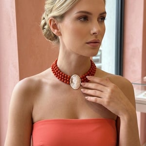 May include: A coral bead necklace with a cameo pendant. The necklace has three strands of round, red coral beads. The pendant is oval-shaped with a white cameo carving. The woman in the image is wearing a coral-coloured strapless top.