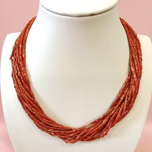 Pode incluir: Um colar de v&aacute;rias voltas composto por pequenas contas cil&iacute;ndricas, vermelho-alaranjadas. O colar &eacute; exibido em um busto de manequim branco sobre um fundo rosa suave. As contas s&atilde;o fortemente amarradas, criando um efeito texturizado e em camadas.