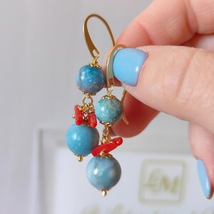 Puede incluir: Pendientes colgantes dorados con tres cuentas de piedra azul-verdosa y peque&ntilde;os detalles de coral rojo. Cierre de gancho dorado. Un accesorio &uacute;nico.