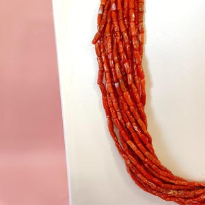 Pode incluir: Um vibrante colar de coral vermelho, composto por v&aacute;rios fios de pequenas contas cil&iacute;ndricas. As contas t&ecirc;m uma forma e textura natural e irregular, com varia&ccedil;&otilde;es sutis de cor. O colar &eacute; exibido em um fundo branco, com um fundo rosa.