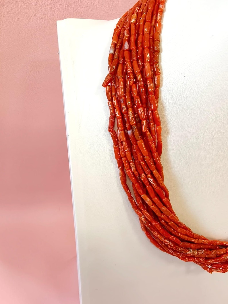 Pode incluir: Um vibrante colar de coral vermelho, composto por v&aacute;rios fios de pequenas contas cil&iacute;ndricas. As contas t&ecirc;m uma forma e textura natural e irregular, com varia&ccedil;&otilde;es sutis de cor. O colar &eacute; exibido em um fundo branco, com um fundo rosa.
