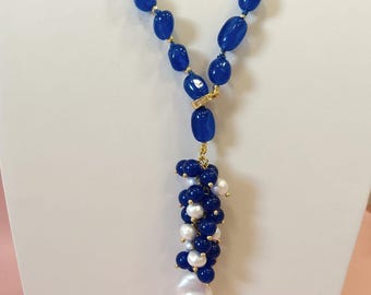 May include: A blue beaded necklace with a gold clasp and accents. The necklace features large, oval-shaped blue beads and a cluster of smaller blue and white beads, culminating in a large, irregular white pearl pendant.