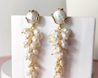 Puede incluir: Un par de pendientes elegantes con una perla blanca grande e irregular en la parte superior, conectada a una cascada de perlas más pequeñas y nacaradas. Los pendientes tienen detalles dorados y se muestran sobre un fondo blanco.