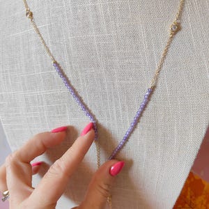Op de afbeelding: Een delicate gouden ketting met een Y-vormig ontwerp. De ketting heeft kleine, lichtpaarse kralen en heldere accentstenen. De ketting is dun en de ketting wordt gepresenteerd op een witte mannequin.