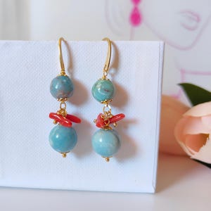 Puede incluir: Pendientes colgantes con ganchos dorados, cuentas de color turquesa y detalles de coral rojo. Los pendientes tienen un dise&ntilde;o en capas con una peque&ntilde;a cuenta en la parte superior, coral en el medio y una cuenta m&aacute;s grande en la parte inferior. Se muestran sobre una superficie blanca.