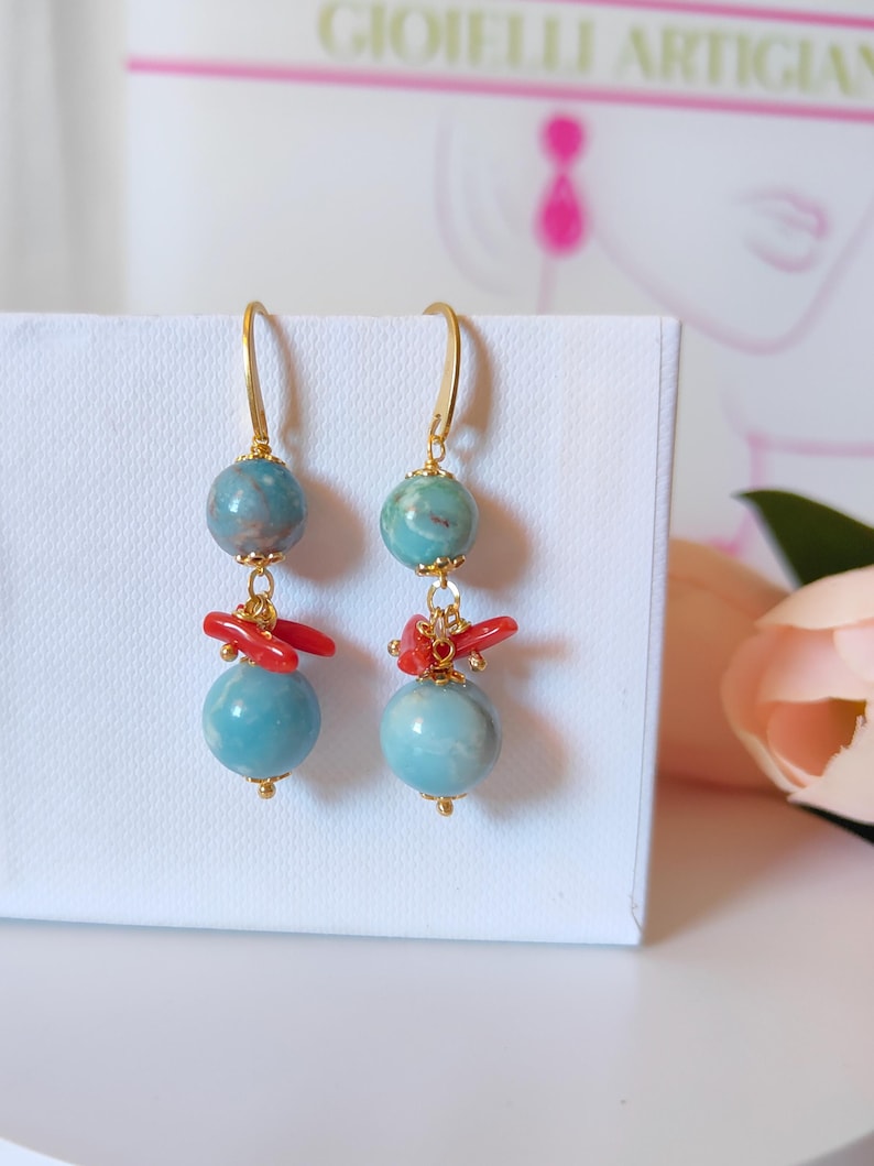 Puede incluir: Pendientes colgantes con ganchos dorados, cuentas de color turquesa y detalles de coral rojo. Los pendientes tienen un dise&ntilde;o en capas con una peque&ntilde;a cuenta en la parte superior, coral en el medio y una cuenta m&aacute;s grande en la parte inferior. Se muestran sobre una superficie blanca.