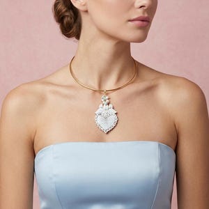 May include: A gold choker necklace featuring a heart-shaped pendant. The pendant is white with a scalloped edge and floral details. The model wears a light blue strapless dress against a pink backdrop.