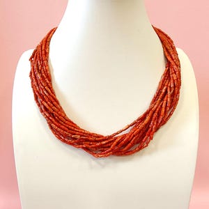 Pode incluir: Um colar de v&aacute;rias voltas composto por pequenas contas vermelho-alaranjadas. O colar &eacute; exibido em um busto de manequim branco contra um fundo rosa suave. As contas parecem estar enfiadas, criando um efeito texturizado e em camadas.