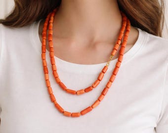 Puede incluir: Un collar de doble hebra con cuentas cilíndricas de color naranja. Las cuentas son uniformes en forma y tamaño, ensartadas para crear dos capas distintas que descansan sobre el cuello. Un pequeño cierre de metal dorado une las hebras.