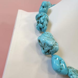 Puede incluir: Un collar compuesto por piedras de turquesa grandes, pulidas e irregulares que presentan vetas de matriz de color marr&oacute;n oscuro. Las piedras est&aacute;n ensartadas en una secuencia continua, mostrando un color azul cielo vibrante y un acabado liso y brillante.