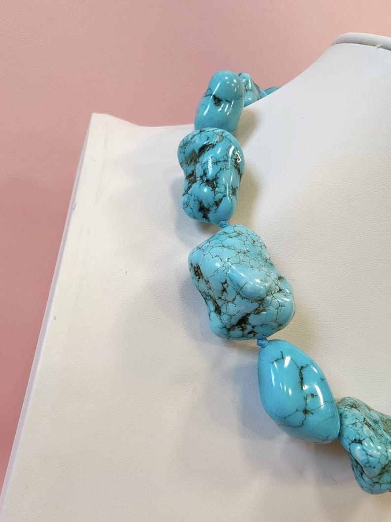 Puede incluir: Un collar compuesto por piedras de turquesa grandes, pulidas e irregulares que presentan vetas de matriz de color marr&oacute;n oscuro. Las piedras est&aacute;n ensartadas en una secuencia continua, mostrando un color azul cielo vibrante y un acabado liso y brillante.