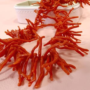 May include: A striking coral-red necklace with a branching, organic design. The necklace is displayed on a light pink surface, with a white jewelry box in the background. The coral-like structure is detailed and eye-catching, suggesting a natural or nautical theme.
