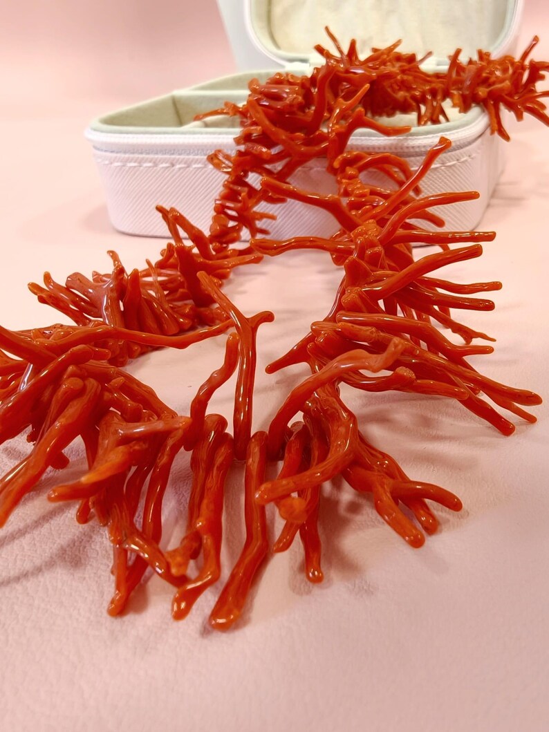 May include: A striking coral-red necklace with a branching, organic design. The necklace is displayed on a light pink surface, with a white jewelry box in the background. The coral-like structure is detailed and eye-catching, suggesting a natural or nautical theme.