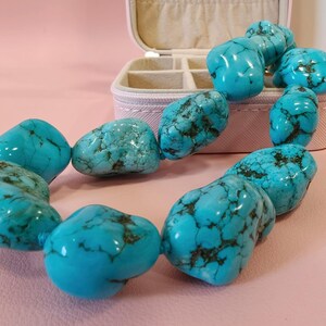 Puede incluir: Un collar compuesto por cuentas de turquesa grandes, pulidas e irregulares con vetas de color marr&oacute;n oscuro. Las cuentas est&aacute;n ensartadas en un bucle continuo, mostrando un color azul vibrante con texturas y patrones de piedra natural.