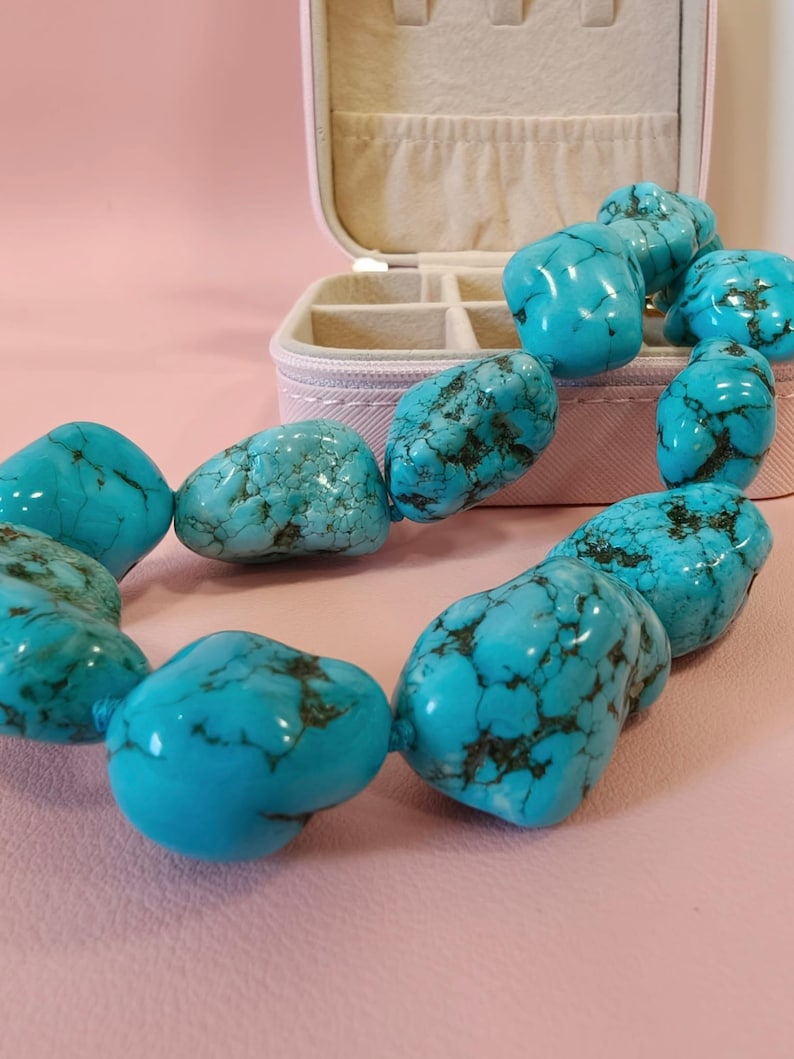 Puede incluir: Un collar compuesto por cuentas de turquesa grandes, pulidas e irregulares con vetas de color marr&oacute;n oscuro. Las cuentas est&aacute;n ensartadas en un bucle continuo, mostrando un color azul vibrante con texturas y patrones de piedra natural.