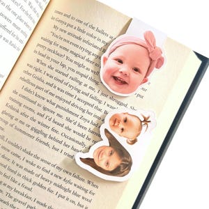 May include: A magnetic bookmark featuring three distinct die-cut portraits of children. The top portrait shows a smiling child wearing a pink headband, while the two smaller portraits below depict children with different hairstyles, all set against a book page.