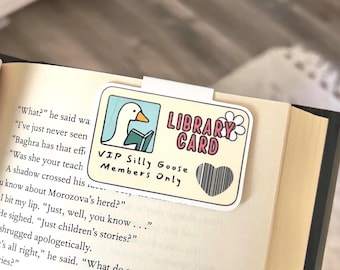 Puede incluir: Un marcapáginas rectangular con un ganso de dibujos animados leyendo un libro, con la etiqueta "Library Card". La tarjeta tiene un corazón y el texto "VIP Silly Goose Members Only". El marcapáginas está sujeto a las páginas de un libro.