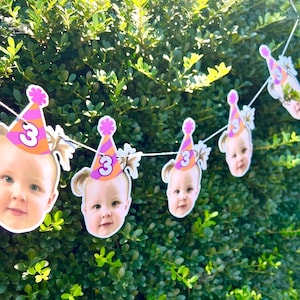 May include: A party banner featuring repeating cutouts of a child's face wearing a pink and orange striped party hat with the number 3 printed on the front. The cutouts are attached to a thin white string and displayed in a repeating sequence.