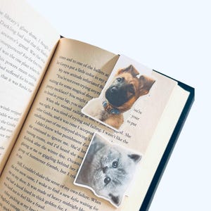 May include: Two pet-themed bookmarks featuring a German Shepherd puppy and a gray cat, placed in an open book. The bookmarks are rectangular with images of the animals. The book has text on the pages.