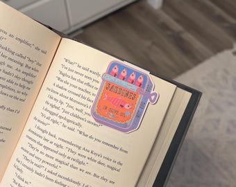 May include: A colorful bookmark featuring a sardine can design with the text "Sardines in Olive Oil." The bookmark is placed on the pages of a book, with the text of the book visible.