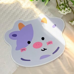 May include: A light gray mousepad with a cartoon cow design. The cow has a purple head, orange horns, a pink nose, and black eyes. The mousepad has a white border and is designed for computer use.