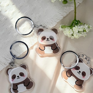 May include: Three panda bear keychains. Each keychain features a clear acrylic panda bear design with brown and white coloring, a smiling face, and a silver keyring. The keychains are displayed on a white surface with a floral accent.