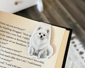 May include: A white Pomeranian dog bookmark is placed in a book. The bookmark shows a detailed illustration of the fluffy dog with a smiling expression. The book has aged pages and black binding.