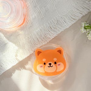 May include: A clear, round silicone lid featuring an orange Shiba Inu dog design. The lid has a cute cartoon face with black eyes, a black nose, and pink cheeks. The lid is designed to fit over a container.