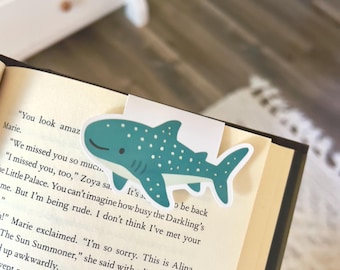 Puede incluir: Un marcapáginas de tiburón ballena turquesa y blanco se coloca en las páginas de un libro abierto. El marcapáginas tiene un diseño de dibujos animados con puntos blancos y una cara sonriente. Las páginas del libro tienen texto impreso.