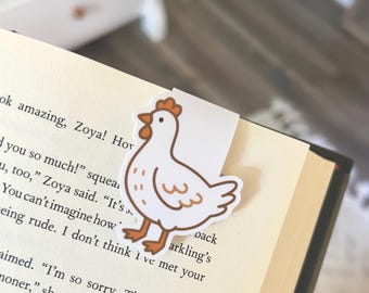 Puede incluir: Un marcapáginas de pollo blanco con detalles marrones, colocado en las páginas de un libro. El marcapáginas presenta un diseño de pollo de dibujos animados con cresta y patas rojas. El texto del libro es parcialmente visible.