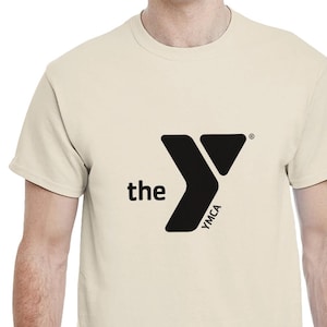 May include: Cream-colored t-shirt featuring the YMCA logo in black. The logo includes the word "the" to the left of a stylized "Y" shape, with "YMCA" printed vertically within the design. The shirt is short-sleeved.
