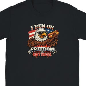Puede incluir: Una camiseta negra de manga corta con el gráfico de un águila calva con gafas de sol, sosteniendo un perrito caliente frente a una bandera estadounidense. El texto dice I RUN ON FREEDOM & HOT DOGS, con una espátula y un tenedor ilustrados.