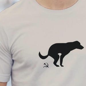 Puede incluir: Camiseta gris claro con una silueta negra de un perro defecando, con un símbolo de la hoz y el martillo debajo. El perro está representado de perfil. La camiseta tiene mangas cortas y cuello redondo.