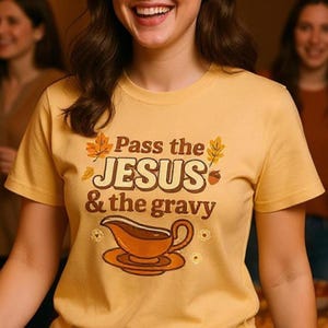 Puede incluir: Camiseta de color amarillo mostaza con la frase "Pass the JESUS & the gravy" en letras marrones. El diseño incluye una ilustración de una salsera y detalles otoñales. La camiseta es de manga corta.