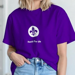 Peut inclure: Un t-shirt violet avec un motif de fleur de lys blanc dans un cercle blanc. Le texte "Ready for life" est imprim&eacute; en dessous. Le t-shirt est port&eacute; avec un jean bleu.