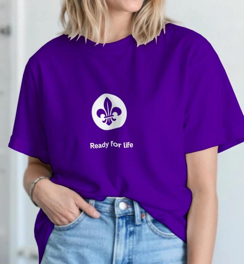 Peut inclure: Un t-shirt violet avec un motif de fleur de lys blanc dans un cercle blanc. Le texte "Ready for life" est imprim&eacute; en dessous. Le t-shirt est port&eacute; avec un jean bleu.