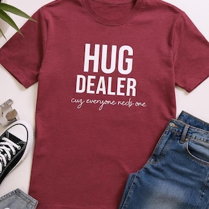 May include: A maroon t-shirt with the words "HUG DEALER" in large white block letters. Below the text is the phrase "cuz everyone needs one" in a cursive font. Blue jeans and a black and white sneaker are also visible.
