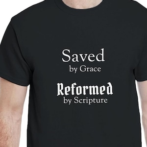 Puede incluir: Camiseta negra con texto blanco. El texto dice "Saved by Grace" encima de "Reformed by Scripture". La camiseta es de cuello redondo y parece estar hecha de algodón o un material similar.