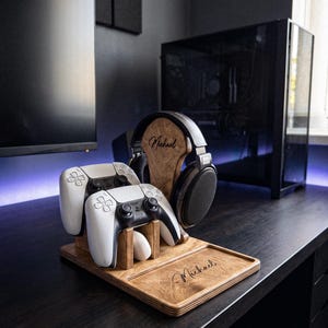 Peut inclure: Un support de jeu en bois comprenant un porte-casque vertical et une base avec des emplacements pour deux manettes blanches. Le bois est teinté d'un brun moyen et porte le nom Michael gravé sur le repose-casque et sur la base.