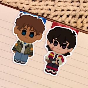May include: Two cartoon character stickers, one with brown hair and a green jacket, the other with black hair and a red and black outfit. They are placed on a lined notebook page.