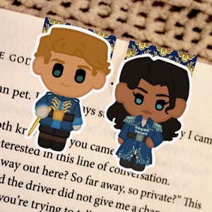 Puede incluir: Dos marcadores de estilo caricatura con personajes de piel clara y oscura. Los marcadores están colocados en un libro, con el texto "THE GOD" visible. Los personajes visten trajes azules.