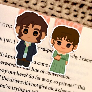 Op de afbeelding: Twee bladwijzers met stripfiguren op een pagina uit een boek. Eén bladwijzer toont een man in een donkerblauwe jas en de andere een vrouw in een groene jurk. De tekst op de pagina luidt "THE GOD" en "why I came".