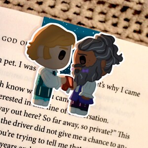 May include: A bookmark featuring two cartoon figures, one with blonde hair and the other with a beard, holding a red object. The bookmark is placed on a book with the text "THE GOD OF" visible.