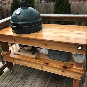 May include: A wooden outdoor grilling station with a dark green ceramic grill on top. The station has two levels with storage and a bottle opener. The wood has a natural finish and is on a wooden deck.