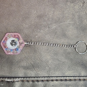 May include: A pink hexagonal keyring with a large googly eye and a silver chain. The keyring is filled with small, colourful beads. The chain connects to a silver ring.