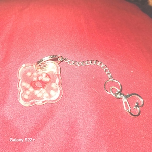 Könnte beinhalten: Ein klarer, vierblättriger Kleeblatt-Schlüsselanhänger mit roter Mitte und weißen Akzenten. Der Schlüsselanhänger ist an einer silbernen Kette und einem herzförmigen Verschluss befestigt. Der Hintergrund ist einfarbig rot.