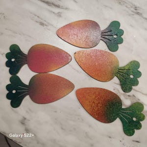 May include: Five wooden carrot-shaped decorations with a gradient of orange, red, and yellow hues. Each carrot has a green leafy top with detailed vein lines. The decorations are arranged on a white and gray marble surface.