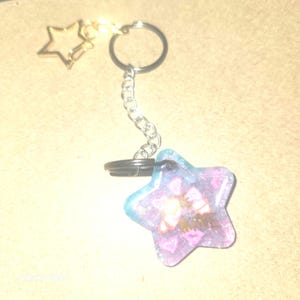 Può includere: Un portachiavi a forma di stella con un ciondolo a forma di stella dorata e una catena argentata. Il ciondolo principale a forma di stella è blu e viola con un disegno e la parola "KISS" in oro. Il portachiavi è attaccato a un portachiavi argentato.