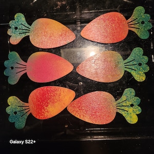 May include: Six wooden carrot cutouts with orange, red, and yellow gradients. The carrot tops are green and blue. The cutouts are arranged in two rows of three on a dark surface. The image is taken with a Galaxy S22+.